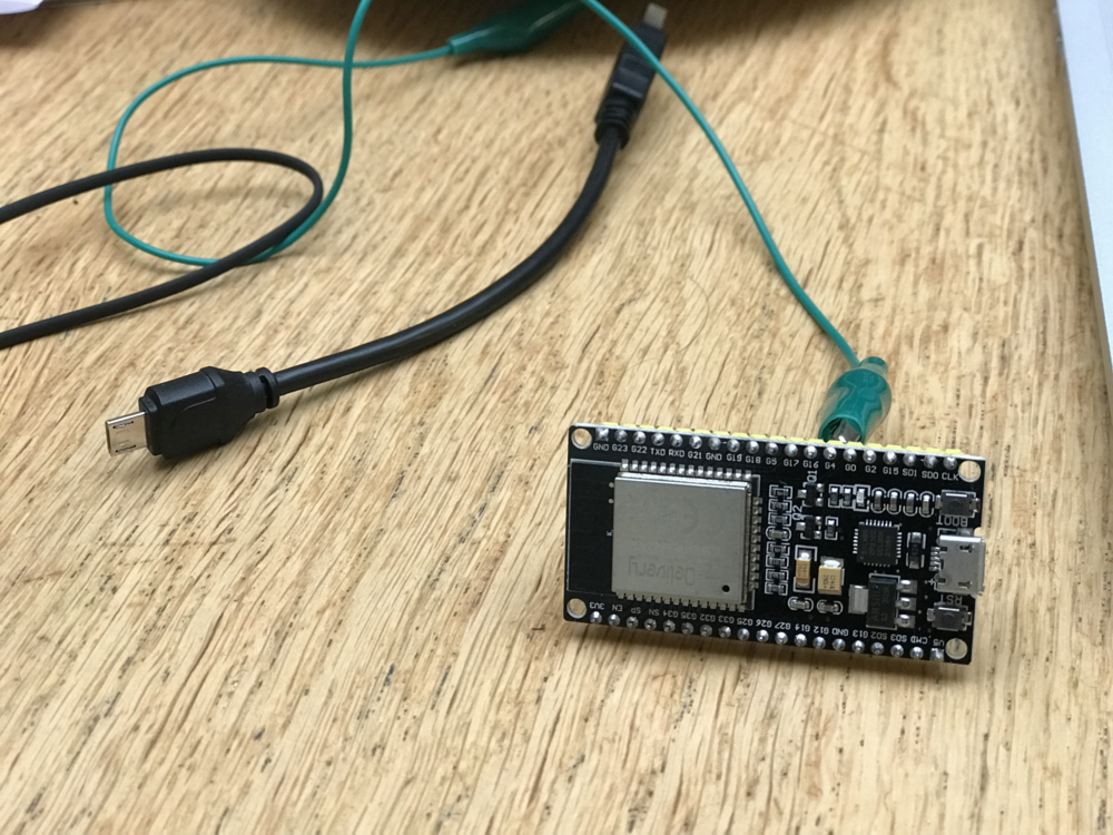 Alligator clip attached to the G0 of a esp32 microcontroller on a table with cables in the background